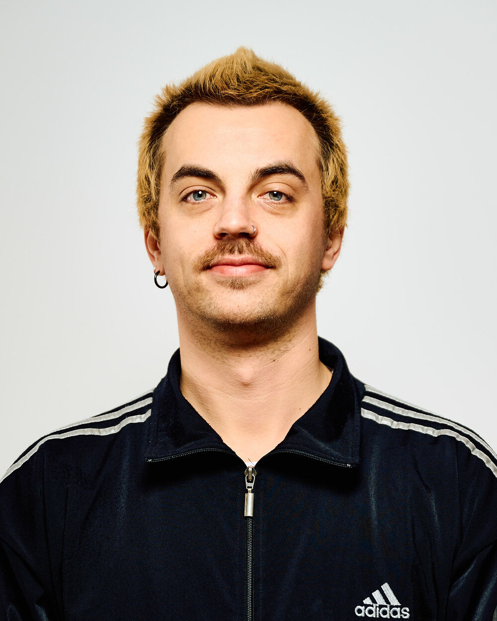 Dieses Bild zeigt Jan. Er hat kurze blonde Haare, die vorne etwas hochgegelt sind. Er hat einen Nasenring und im rechten Ohr einen Ohrring. Er trägt eine dunkelblaue sportliche Jacke. 