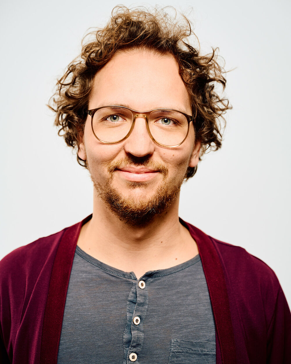 Dieses Bild zeigt Christoph. Er hat kurze gelockte Haare und einen kurzen Bart. Er trägt eine braun gerahmte Brille und ein graues Shirt über dem er eine dunkel rote Jacke angezogen hat. 