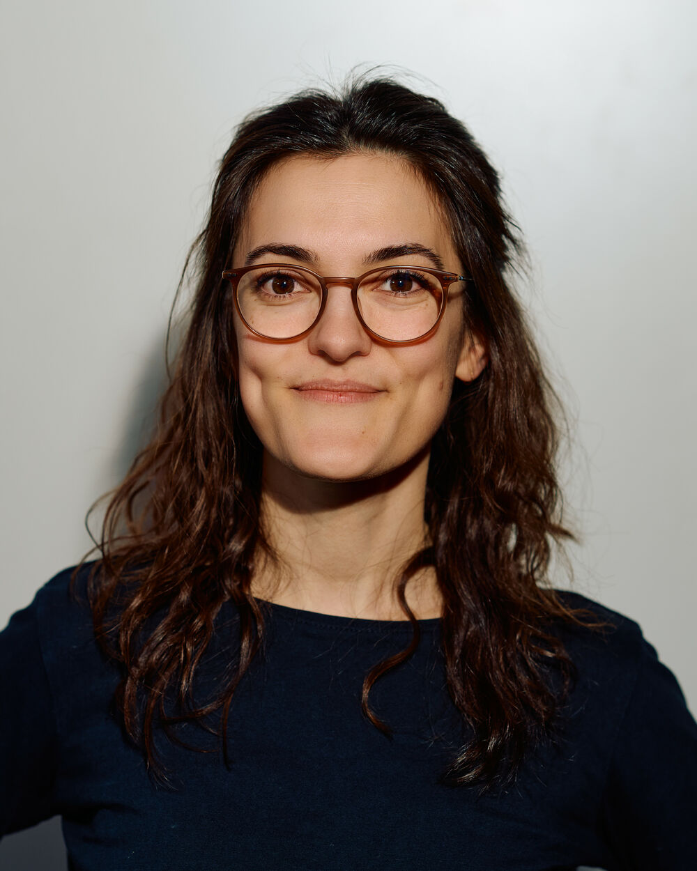 Eine Frau mit Brille, langen dunklen Haaren und einem leichten Lächeln, die ein schwarzes Oberteil trägt, steht vor einem neutralen Hintergrund. Das Bild hat einen warmen, zugänglichen Ton.