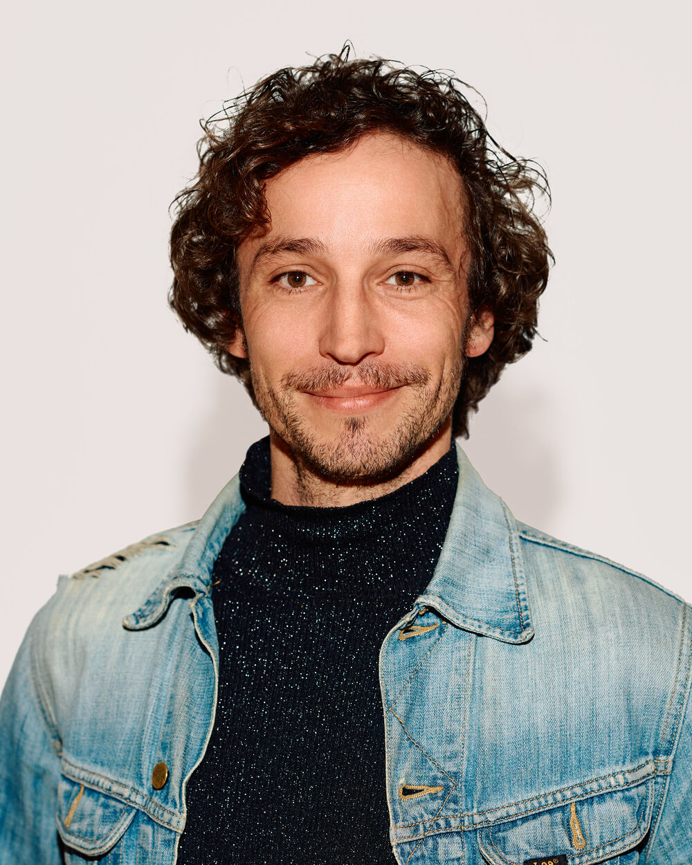 Porträt eines lächelnden Mannes mit braunen Locken und Dreitagebart, der einen dunklen Rollkragenpullover und eine hellblaue Jeansjacke vor hellem Hintergrund trägt.