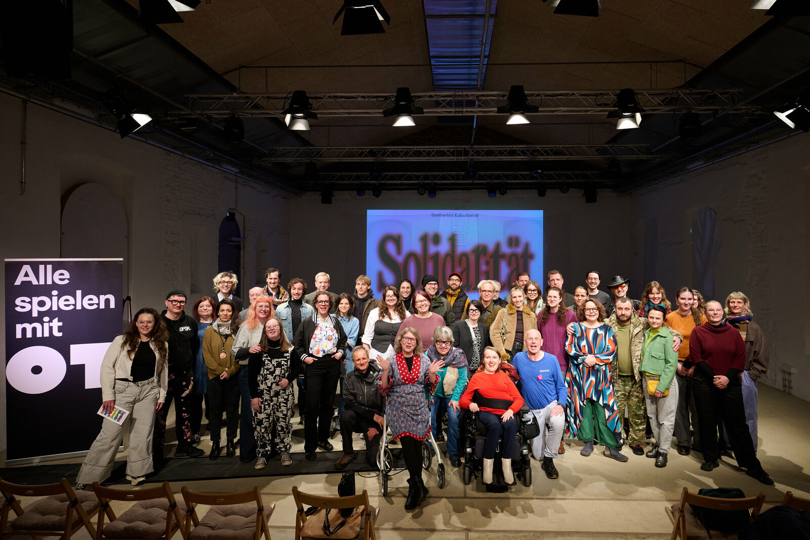 Ein großes Gruppenfoto mit über 30 lächelnden Personen, die zusammen auf einer Bühne in einem Veranstaltungssaal stehen. Links im Vordergrund steht ein Banner mit der Aufschrift „Alle spielen mit 01“, und im Hintergrund ist auf einer Leinwand der Schriftzug „Solidarität“ zu sehen. Die Gruppe ist divers und inklusiv, einige Personen sitzen im Rollstuhl. Die Atmosphäre ist fröhlich und gemeinschaftlich. Der Raum ist mit professionellen Bühnenlichtern beleuchtet.