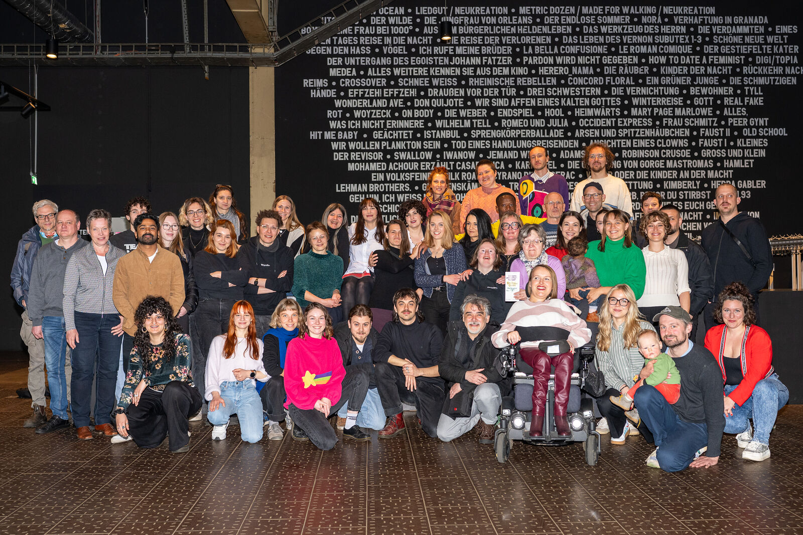 Eine bunt gemischte Gruppe von Menschen, die lächelnd für ein Foto in einem Theater posieren. An der Wand hinter ihnen steht der Titel eines Theaterstücks. Warme, jubelnde Atmosphäre.