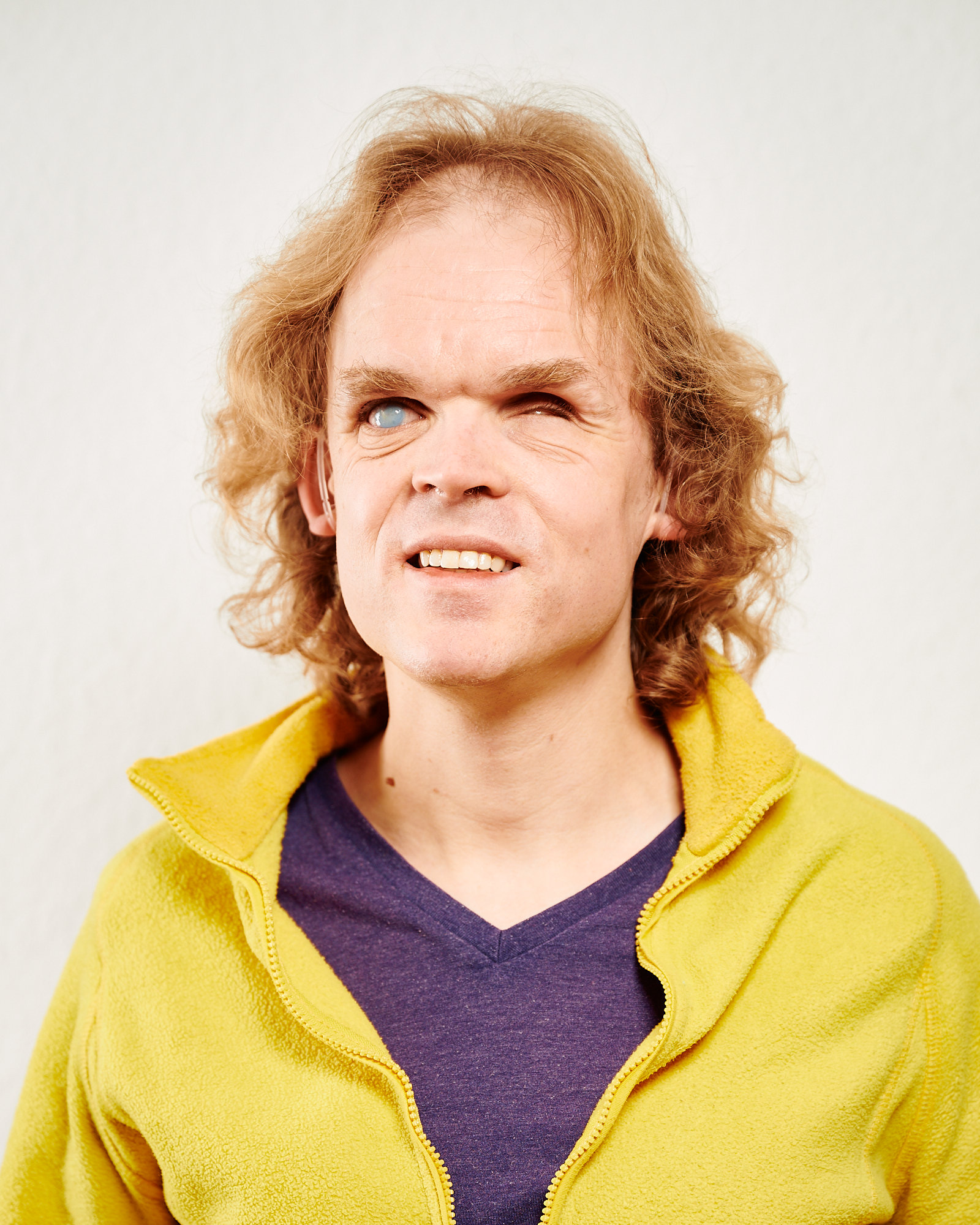 Dieses Bild zeigt Arne. Er hat lange, hellbraune, lockige Haare. Er trägt ein lila T-Shirt und einen gelben Fleece-Pullover. 